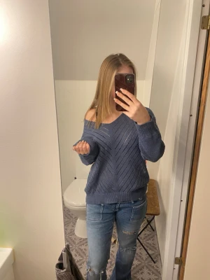 Blå stickad tröja med v-ringning - Mysig blå stickad tröja med v-ringning och avslappnad passform. Tröjan har ett diskret hålmönster i diagonala ränder och breda muddar vid ärmslut och nederkant. Perfekt att slänga på sig över ett linne eller t-shirt.