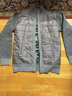 Grå cardiganjacka med stickade ärmar - Snygg grå cardiganjacka med stickade ärmar och rygg samt vadderad framsida. Jackan har ribbstickad krage, muddar och nederkant samt dragkedja framtill. Inga fickor. Perfekt för lager-på-lager-stil.Helt ny