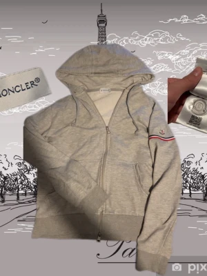 Grå hoodie från Moncler med dragkedja - Snygg grå hoodie från Moncler med hel dragkedja, huva och snörning. Klassisk Moncler-logga på ärmen och en diskret röd, vit och blå randdetalj. Tillverkad i mjuk bomull, perfekt för en chill och stilren look.strl xs / s