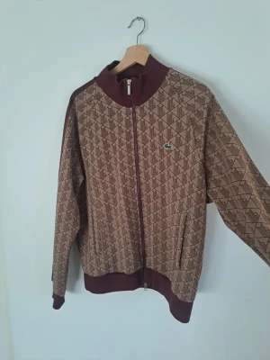 Brun mönstrad ziptröja från Lacoste - Snygg brun ziptröja från Lacoste med grafiskt mönster och klassisk krokodillogga på bröstet. Tröjan har ribbade muddar, hög krage och dragkedja hela vägen. Perfekt för en sportig och trendig look.