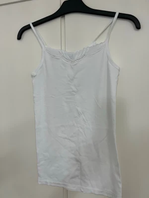 Vit linnetopp med spetsdetalj - Säljer en enkel och fräsch vit linnetopp med tunna axelband och snygg spetsdetalj vid urringningen. Perfekt att styla lager på lager eller bära som den är. Materialet känns mjukt och lätt.