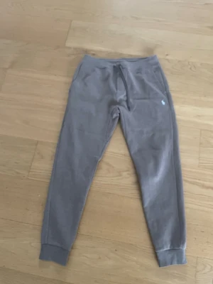 Gråa mjukisbyxor från Polo Ralph Lauren - Säljer ett par gråa mjukisbyxor från Polo Ralph Lauren med dragsko i midjan och ribbade muddar vid bensluten. Byxorna har fickor på sidorna och den klassiska vita Polo-loggan broderad på vänster sida. Perfekta för chill dagar eller när du vill vara bekväm.