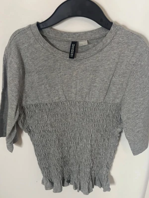 Grå smockad topp från H&M  - Grå topp från H&M Divided med smockad midja och volangkant nedtill. Perfekt till jeans eller kjol❤️