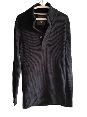 Svart ribbstickad tröja - Svart ribbstickad tröja med hög krage och två knappar vid halsen. Långärmad modell med breda muddar vid ärmslut och nederkant. Perfekt för lager-på-lager och enkel att matcha med olika outfits.