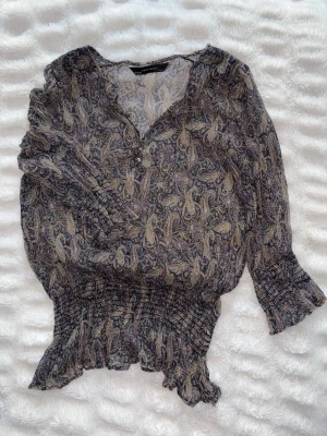 Mönstrad blus från zara - Säljer en tunn, mönstrad blus från zara med paisleymönster. Blusen har trekvarts ärmar med smockdetaljer vid ärmslut och midja samt v-ringning med knappar. Perfekt för en trendig och bohemisk look. Tråd lös vid ena armen se bild