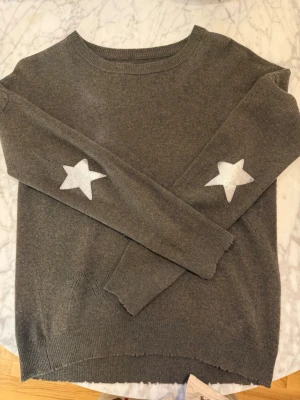 Zadig & Voltaire Grön stickad tröja med sjärnor - Mysig grå stickad tröja från Zadig & Voltaire med silvriga stjärnor på armbågarna. Tröjan har rund halsringning, långa ärmar och ribbade muddar. Den är i väldigt fint skick och knappt använd. 