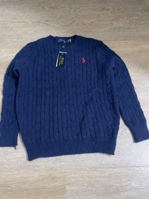 Mörkblå kabelstickad tröja Polo Ralph Lauren - Mörkblå kabelstickad tröja från Polo Ralph Lauren med rund halsringning och röd broderad logga på bröstet. Tröjan har ribbstickade muddar vid ärmslut och nederkant. Perfekt för en stilren och klassisk look.