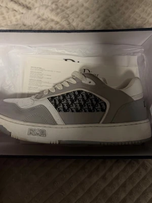 Dior B23 grå och vita sneakers - Snygga Dior B23 sneakers i grått och vitt med ikoniskt Dior-mönster på sidan. Skorna har låga skaft, vita snören och detaljer i läder och textil. Perfekta för dig som vill ha en exklusiv och trendig look.