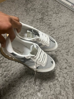 Valentino Rockrunner sneakers beige/grå - Snygga Valentino Rockrunner sneakers i beige, vitt och grått med kamouflagemönster. Skorna har vita snören, mesh- och mockadetaljer samt den ikoniska nitar på hälen. Sulan är beige med gummidetaljer och logga på plösen.