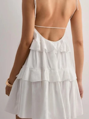 Vit volangklänning med öppen rygg - Söt vit klänning med tunna axelband och öppen rygg. Klänningen har flera lager volanger som ger en luftig och lekfull känsla. Perfekt för varma dagar och somriga tillfällen. Materialet ser ut att vara lätt och skönt.