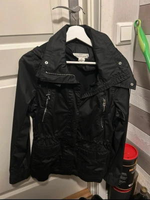 Svart vindjacka med dragkedjor - Snygg svart vindjacka från H&M med hög krage och dragkedjor på bröstfickorna. Jackan har justerbar midja och två stora fickor framtill. Perfekt för blåsiga dagar och enkel att matcha med olika outfits.