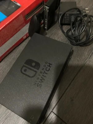 Nintendo Switch med neonröda och blå Joy-Con - Nintendo Switch spelkonsol med neonröda och blå Joy-Con-kontroller. Konsolen har en svart dockningsstation och medföljer laddare. Perfekt för gaming både hemma och på språng. Materialet är mestadels plast och färgerna är svart, röd och blå.