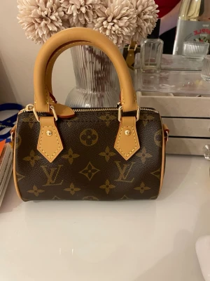 Louis Vuitton Speedy Nano Monogram - Säljer en Louis Vuitton Speedy Nano handväska i klassiskt brunt monogramcanvas med beige läderdetaljer och guldfärgade metalldelar. Väskan har två rundade handtag och dragkedja upptill. Ikonisk och tidlös design som är superpopulär just nu.
