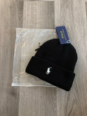 Ribbstickad mössa från Polo Ralph Lauren - Snygg ribbstickad mössa från Polo Ralph Lauren med klassisk uppvikt kant och broderad logga framtill. Finns i både svart och ljusgrått. Perfekt accessoar för att hålla värmen och ge din outfit en sportig touch.