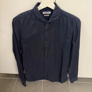 Mörkblå skjorta från J.Lindberg - Stilren mörkblå skjorta från J.Lindberg med klassisk krage och knäppning framtill. Skjortan har lång ärm och en enkel, tidlös design som passar till många olika outfits. Perfekt för dig som gillar en clean och snygg look. Storlek S