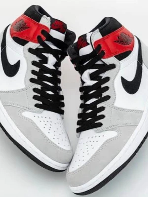 Nike Air Jordan 1 High OG SKORNA FINNS INNE, BILDER SKICKAS PÅ BEGÄRAN - Snygga Nike Air Jordan 1 High OG sneakers med vit bas, grå mockadetaljer, svart swoosh och snörning samt röd accent vid ankeln. Klassisk high-top modell med ikonisk vinge-logga på sidan. Perfekta för dig som gillar streetwear och sportig stil.