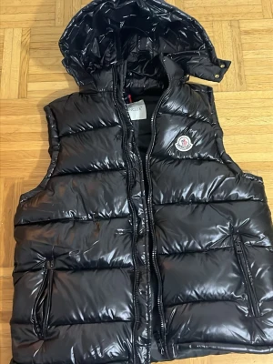 Moncler väst  - Snygg svart dunväst från Moncler med glansig finish och avtagbar luva. Västen har dragkedja framtill, två fickor med dragkedja och Moncler-logga på bröstet. Perfekt för lager-på-lager och streetwear-vibe.