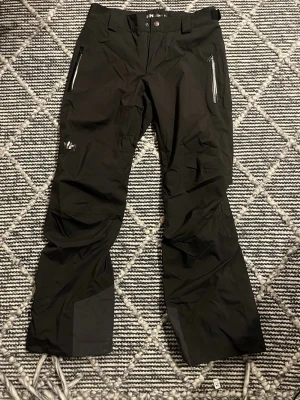  skidbyxor från Helly Hansen - Säljer ett par svarta skidbyxor från Helly Hansen med dragkedjeförsedda fickor framtill och förstärkta partier vid bensluten. Byxorna har bälteshällor och HH-logga på benet. Perfekta för vintersport och outdoor. Använda endast en vecka. Lite vit på baksidan pga förvaring.