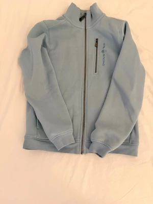 Ljusblå fleecejacka Sail Racing - Ljusblå cardiganjacka från Sail Racing i mjuk fleece. Jackan har hel dragkedja, hög krage och en bröstficka med dragkedja. Diskret logga på bröstet och ribbade muddar vid ärmslut och nederkant. Perfekt för lager-på-lager och chill dagar.