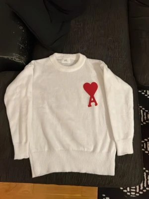 Vit stickad tröja från Ami Paris - Säljer en vit stickad tröja från Ami Paris med rund halsringning och ribbade muddar. På bröstet finns ett stort rött hjärta med bokstaven A, vilket är signaturdetaljen från märket. Tröjan har en avslappnad passform och är perfekt för dig som gillar stilrena plagg med en twist.