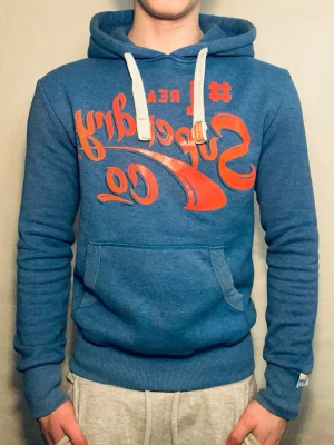 Blå Superdry hoodie med tryck - Snygg blå hoodie från Superdry med stort orange tryck på bröstet och vita dragsnören i huvan. Klassisk känguruficka framtill och ribbade muddar vid ärmar och midja. Perfekt för en avslappnad streetwear-look.