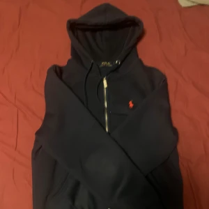 Mörkblå hoodie från Polo Ralph Lauren - Säljer en Mörkblå hoodie från Polo Ralph Lauren med dragkedja framtill och klassisk röd logga broderad på bröstet. Hoodien har huva med snörning. Perfekt för en avslappnad och stilren look. Pris kan diskuteras, mer bilder kan skickas, ställ gärna frågor vid osäkerhet. Har använd den väldigt få gånger, definitivt under 10.