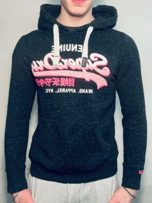 Svart Superdry hoodie med tryck - Säljer en svart hoodie från Superdry med vitt och rosa tryck på bröstet. Hoodien har vita snören, känguruficka och en klassisk huva. Materialet är mjukt och skönt, perfekt för chill dagar. Superdry-logga även på ärmen.