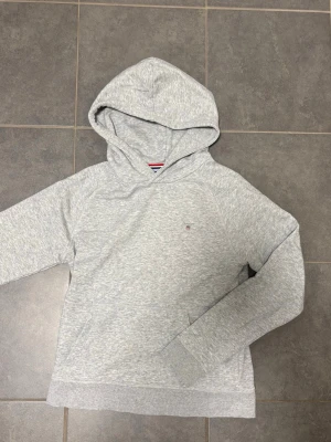Ljusgrå hoodie från Gant  - Snygg ljusgrå hoodie från Gant med liten broderad logga på bröstet. Klassisk modell med huva och känguruficka framtill. Mjuk bomullsblandning och ribbade muddar vid ärmslut och nederkant. Perfekt för en avslappnad och stilren look.