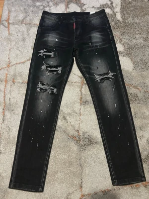 Svarta slitna jeans Dsquared2 - Säljer ett par svarta skinny jeans från Dsquared2 med slitningar och hål på framsidan, samt vita färgstänk över hela byxan. Baksidan har broderade blommor och blå D2-logga ovanför ena bakfickan. Snygg streetstil och trendig look.