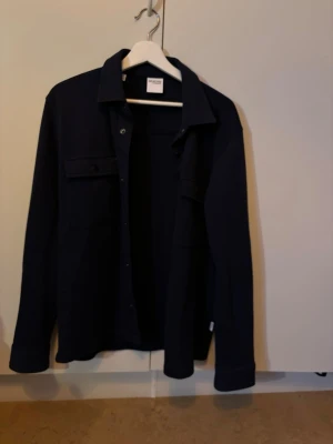 Mörkblå overshirt från Selected Homme - Stilren mörkblå overshirt från Selected Homme med klassisk krage och tryckknappar framtill. Två bröstfickor med lock och långa ärmar. Perfekt lager-på-lager-plagg i mjukt material som ger en avslappnad men snygg look.