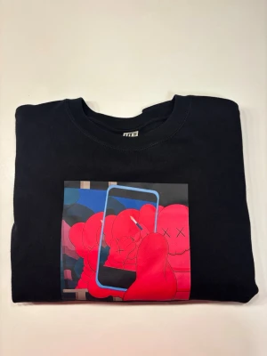 Svart sweatshirt med KAWS-print - Cool svart sweatshirt från Uniqlo UT med färgstarkt KAWS-motiv på bröstet. Trycket har röda figurer och blå detaljer, vilket ger en unik streetwear-känsla. Tröjan har rund hals och är tillverkad i mjuk bomull. Perfekt för dig som gillar konst och popkultur.