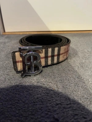 Två sidigt burrbery bälte  - Snyggt bälte från Burberry med det ikoniska beige, röda och svarta rutmönstret. Bältet har ett stort metallspänne med Burberrys TB-logga i silverfärg. Perfekt accessoar för att lyfta din outfit med en lyxig touch.
