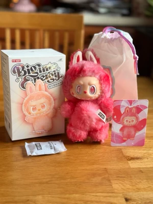 Pop Mart The Monsters Labubu Big into Energy Love - Söt samlardocka från Pop Mart, Bigmine-serien, klädd i fluffig rosa dräkt med kaninöron och stora glittriga ögon. Dockan är gjord av plast och mjukt tyg. Medföljer originalförpackning, samlarkort och tygpåse.