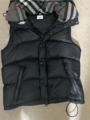 Svart Burberry dunväst med huva - Snygg svart dunväst med dragkedja och knappar framtill. Västen har två fickor med dragkedja och en avtagbar huva med rutigt foder i grått, vitt och rött. Perfekt för lager på lager och streetwear-stil.