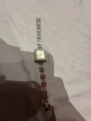 Anne Klein fyrkantig silverklocka - Stilren klocka från Anne Klein med fyrkantig urtavla i silver och länkarmband i både silver och brunt. Klassisk design med minimalistisk urtavla och diskreta siffror. Perfekt accessoar för dig som gillar en tidlös look. Funkar ej!