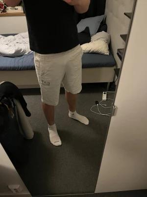Vita EA7 shorts med tryck - Säljer ett par vita shorts från EA7 med svart logotyptryck på sidan och framsidan. Snygg och sportig look som funkar till många outfits.