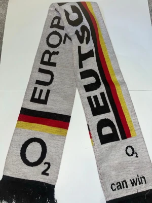 Grå halsduk med tysk flagga - Snygg grå halsduk med texten 'EUROPA', 'DEUTSCH' och detaljer i svart, rött och gult som påminner om tyska flaggan. Halsduken har fransar i ändarna och är stickad i mjukt material. Perfekt för att visa ditt stöd för Tyskland.