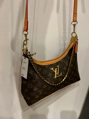 Louis Vuitton monogram axelväska - Snygg axelväska från Louis Vuitton med klassiskt monogrammönster i brunt och beige. Väskan har guldfärgad kedja, justerbar axelrem med röda detaljer och LV-logga framtill. Tillverkad i skinn med exklusiv känsla och fina detaljer.