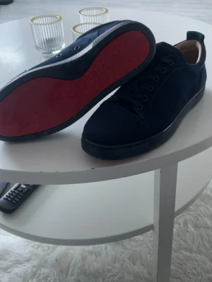Mörkblå sneakers från Christian Louboutin - Snygga mörkblå sneakers från Christian Louboutin i mocka med klassisk röd sula och svart snörning. Diskret broderad logga på hälen och låg profil gör dem perfekta för en clean och stilren look.