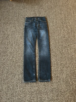 Mörkblå jeans från Nudie Jeans - Snygga mörkblå jeans från Nudie Jeans med klassisk femficksmodell och kontrastsömmar. Jeansen har en rak passform och är tillverkade i slitstarkt denimtyg. Perfekta för dig som gillar en tidlös och stilren look. W25 L32