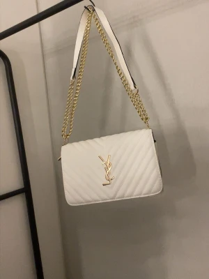 Vit YSL handväska med kedja - Snygg vit handväska från YSL med quiltat mönster och guldiga detaljer. Väskan har en axelrem i vitt läder kombinerat med en guldig kedja. Klassisk rektangulär form och ikonisk YSL-logga framtill. Perfekt för dig som vill ha en trendig och lyxig accessoar.