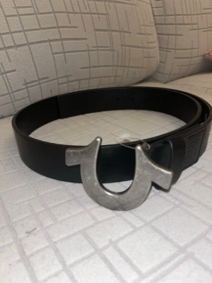 Svart skärp från True Religion - Snyggt svart skärp från True Religion med bred rem i skinn och ikoniskt stort silverspänne i form av varumärkets logga. Perfekt accessoar för att lyfta din outfit med en cool detalj.