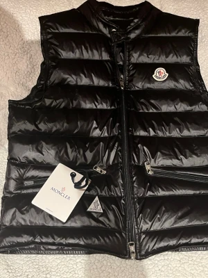 Svart dunväst från Moncler - Snygg svart dunväst från Moncler med glansig finish och klassisk logotyp på bröstet. Västen har två dragkedjeförsedda fickor framtill och hög krage. Perfekt för lager-på-lager och streetwear-stil. Pris går diskutera.