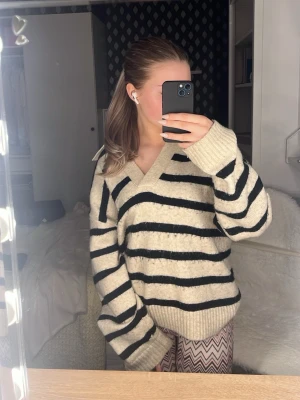 Beige stickad tröja med svarta ränder - Mysig stickad tröja i beige med breda svarta ränder och v-ringad hals. Tröjan har en avslappnad passform och långa ärmar med ribbade muddar. Perfekt för lager-på-lager och chill dagar. Tröjan är väldigt stor i sin storlek.