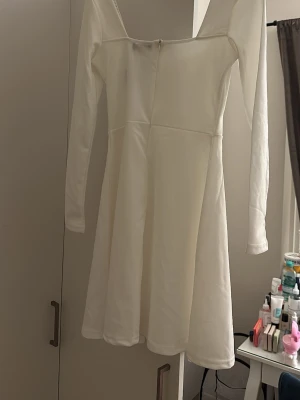 Vit långärmad klänning med pärldetalj - Snygg vit klänning med lång ärm och pärlor längs urringningen. Klänningen har en lätt utsvängd kjol och markerad midja, vilket ger en feminin siluett. Perfekt för dig som vill ha en stilren och elegant look.