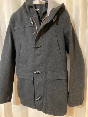 Pull and Bear str small - Nyskick  - Stilren mörkgrå skepparkavaj från Pull and Bear  med huva och klassiska träknappar. Jackan har två stora fickor framtill och är tillverkad i ullblandning, storlek Small 