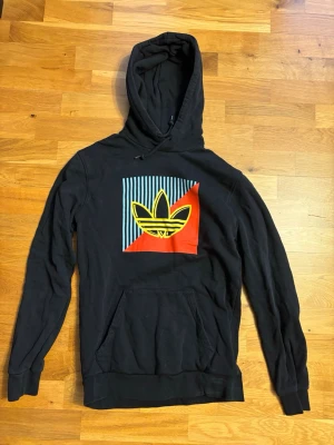 Adidas Hoodie | svart med tryck   - Svart hoodie från Adidas med stor färgglad logga på bröstet i gult, rött och blått. Klassisk huva med dragsko och stor magficka framtill. Tillverkad i mjukt material, perfekt för en avslappnad streetwear-look.