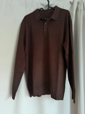 Brun långärmad pikétröja från nuvéclothing - Snygg brun långärmad pikétröja med krage och knappar framtill. Tröjan har en klassisk passform och ribbade muddar vid ärmslut och nederkant. Perfekt för en stilren och avslappnad look.