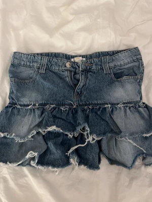 Blå jeanskjol med volanger - Säljer en blå jeanskjol med volanger och råa kanter. Kjolen har klassisk femficksmodell, bälteshällor och knäppning fram.