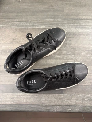 Svarta sneakers från PACE - Stilrena svarta sneakers från PACE med klassisk låg profil och vita sulor. Skorna har svarta skosnören och är tillverkade i ett slätt material som ger en clean look. Perfekta för dig som gillar minimalistisk stil och vill ha ett par enkla, snygga sneakers. OBS inte tvättade på bilden( gör det innan skickar såklart) nypris 799, pris kan diskuteras 
